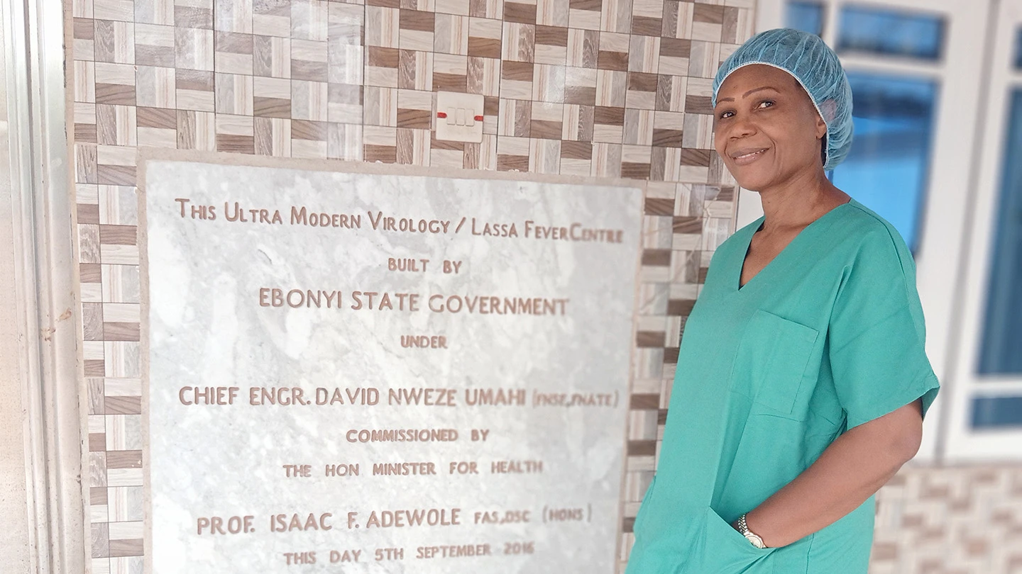 Dr Nnennaya in scrubs standing by plaque of Lassa fever treatment centre