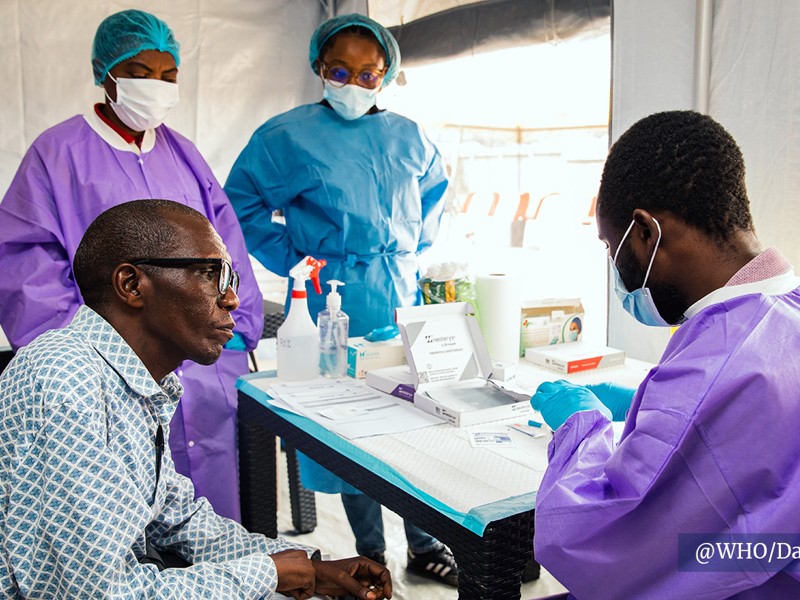 Health workers in protective gowns talk with a seated man at a screening table at a Tumaini trial site, in an image published by CEPI and credited to WHO.